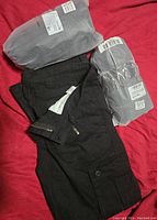 Photo showing three pairs of men's pants. Two pairs are still wrapped in packaging, and one black pair is unwrapped and laid flat.
