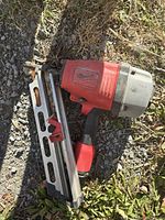 Photo of a Milwaukee nail framing tool lying on grass and dirt ground. Shows red and gray body, metal nail magazine, and handle grip.