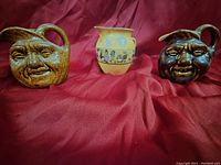 Three pottery items arranged on a red cloth background; includes two brown moon face pitchers and one small yellow jug with cartoon decal band.