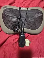 Ogawa electric neck massager placed on red fabric background, showing massager surface with mesh covering, power cord and plug adapter included.