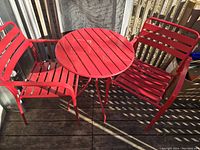 Red metal bistro set shown on wooden deck, includes round table and two matching chairs