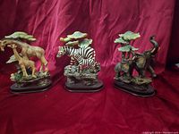 Lot featuring three safari animal figurines including giraffes, zebra, and elephants against a red fabric background.
