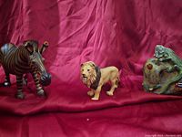 A group photo of three safari animal figurines including a zebra, lion, and hippopotamus on a red fabric background.