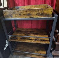 Wooden serving tray on top of black metal frame storage unit with two lower wooden shelves visible.