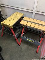 Two Builder's Pal collapsible work benches side by side, showing red metal frames and light wooden tops with holes.