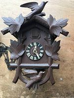 Front photo of a wooden cuckoo clock featuring carved birds and leaves with a black clock face showing Roman numerals