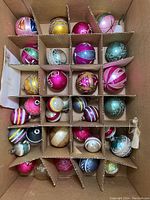 Top-down photo of a box with 30 compartments holding various vintage glass ball ornaments of pink, blue, green, and gold colors with painted and glitter patterns.