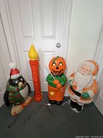 Full view of four vintage blow molds: Christmas penguin, tall orange candle, Halloween pumpkin man, and Santa Claus figure in front of a white door.