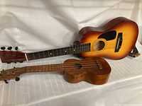 Photo showing both the sunburst First-Act Discovery acoustic guitar and the smaller natural wood Pepe Hawaii ukulele lying side by side on a white cloth.