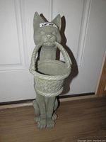 Photo showing the entire gray resin cat statue standing with a woven basket held in its mouth, placed on a wooden floor in front of a white door.