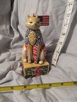 Front view of Jim Shore cat figurine with American flag design, sitting on a decorative base, placed near measuring tapes showing its size.