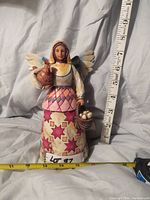 Full front view of the Jim Shore Angel of Faithfulness figurine showing detailed painted patterns, holding hen and basket of eggs, with measuring tape for size reference.