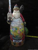 Full front view of the Jim Shore Santa figurine displaying the painted village and church details on the robe, Santa holding a church model, and carrying a green sack and sticks.