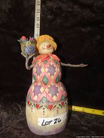 Full view of the Jim Shore snowman figurine showing colorful patchwork design and flower pot in hand against a black background and measuring tape.