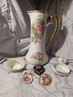 Photo showing Limoges pitcher with floral pink roses design, gold handle and rim, two small dishes, heart shaped trinket box with gold crest, and two pieces of jewelry with figurative painted designs.