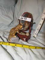 Porcelain cat figurine sitting on a cushioned stool looking into a swivel mirror with vanity table base, measured with tape for scale.