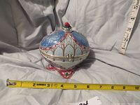 Closed view of decorative egg showing intricate painted enamel and metalwork with measuring tapes indicating size.