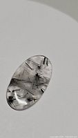 Close-up image of an oval cabochon black rutilated quartz with black tourmaline inclusions lying on a white background, visible needle-like inclusions and polished surface.