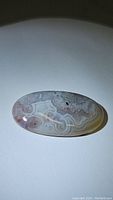 Top view of the oval cabochon cut white Crazy Lace Agate showing the natural swirling patterns in white, light gray, and purple hues with a polished surface.