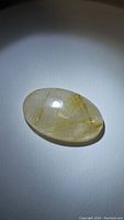 Top-view photo of the transparent oval cabochon featuring golden needle rutile inclusions inside the quartz stone.