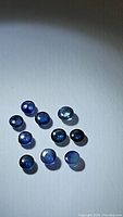 Ten small blue round cabochon cut sapphires arranged on a plain surface with light reflection showing their polished dome shape and blue hues.