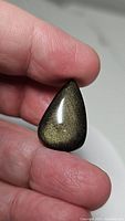 Close-up photo showing the pear-shaped golden sheen obsidian cabochon held between thumb and forefinger against a neutral background. The cabochon is polished with golden sheen reflections visible.
