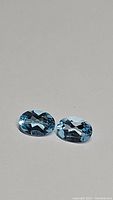 Pair of sky blue natural oval cut topaz gemstones on light grey background showing color and clarity.