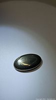 Close-up photo of a natural pyrite oval cabochon gemstone with metallic luster on white background.