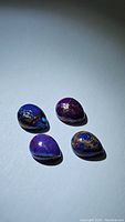 Four pear-shaped purple copper turquoise cabochon gemstones with golden veins positioned on a plain surface under direct light, showing top-down and angled views of the stones.