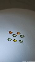 Seven oval cut rainbow sapphires arranged on a white surface, showing color variation including green, yellow, orange, and blue.