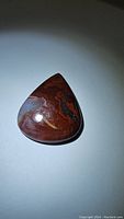 Close-up photo of a polished pear-shaped cabochon red crazy lace agate gemstone showing intricate banded patterns in shades of red, brown, orange, grey and black with a smooth, glossy surface.
