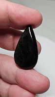 Close-up photo of a pear-shaped cabochon natural obsidian gemstone displaying smooth polished finish and dark base with silver sheen.