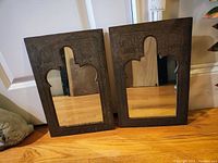 Two carved wood framed mirrors standing on wood floor showing distinctive mirror cutouts and carved detailing around frames.