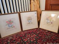 Three framed floral prints resting on a patterned rug against a wall with coats hanging, showing arrangement and overall condition.