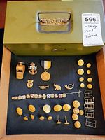 Entire lot displayed inside vintage metal lockbox showing all items including military medal, pins, buttons, whistle, bracelet, buckles.
