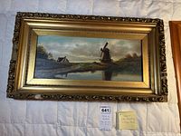 Horizontal oil painting in a gold ornate antique frame on a white surface with lighting showing the windmill and house landscape.