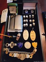 Photo showing all items in the lot on a display surface, including the travel clock, flask, cufflinks in box, bracelet, keychains, pins, whistle, bottle opener, and pipe.