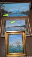 Three vintage oil paintings stacked showing maritime and landscape scenes, varied frames; top one shows mountains, trees and water.