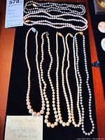Seven strands of vintage faux and freshwater pearl necklaces laid out on black background showing varying pearl sizes and colors with gold clasps.