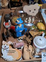 Wide shot showing the entire box of mixed vintage figurines, miniatures, and calendar items with items overlapping, including the Welsh Corgi Calendar, painted eggs, teddy bear head, and teapot.