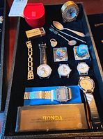 Photo showing three vintage Mickey Mouse watches (two without straps), one with leather strap, Honda digital watch in original case, Adidas watch, Hamilton desk clock, two glass swirl paperweights, three collector spoons, and a metal heart-shaped lock on display.