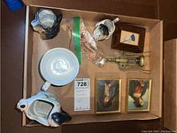 Top view of all items in lot inside a cardboard box: 3 framed prints, 3 animal-shaped pitchers or cups, brass corkscrew, and two other cups or figurines.