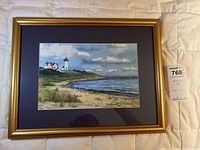 Framed wall art print showing a lighthouse scene with houses, beach, and ocean under partly cloudy sky. Gold frame and blue matte visible.