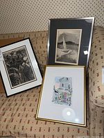 Three framed prints displayed on a sofa showing various artistic scenes including a sailboat, urban scene with a person, and a colorful building front.