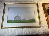 Full view of framed watercolor showing house and smaller structure with grassy foreground and cloudy sky, on a bed surface.