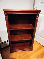 Front view of small wood bookcase showing three shelves and decorative carved columns on the sides. Top shows wear and scratches.