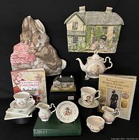 Overall lot image showing Beatrix Potter tea set items arranged with books and music box, including teapot, several creamers, cups, saucers, a small plate, and two fabric tea cozies.