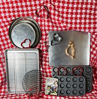 Photo shows four cookie sheets stacked, two wire baking racks, two mini cupcake pans with 24 small cavities each, one aluminum baking tray with metal heart and cat cookie cutters on it, and a small package of butter dragon cookies.