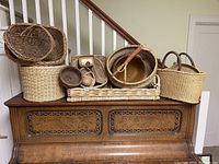10 assorted baskets arranged on top of a wooden piano, showing variety in shape, size, and weave styles. Some have handles, others are basket boxes or trays made from woven natural fibers.