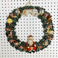 Full view of the 16-inch handmade signed clay Christmas wreath hung on pegboard with holiday motifs including Santa, candy canes, drums, dolls, and presents.